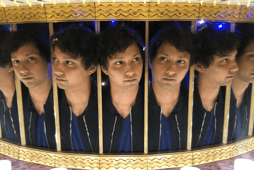 Joey Reyes, wearing a navy blue jacket and a lighter blue v-neck shirt, sticking his head into a mirrored box, so there are numerous reflections to his left and right.